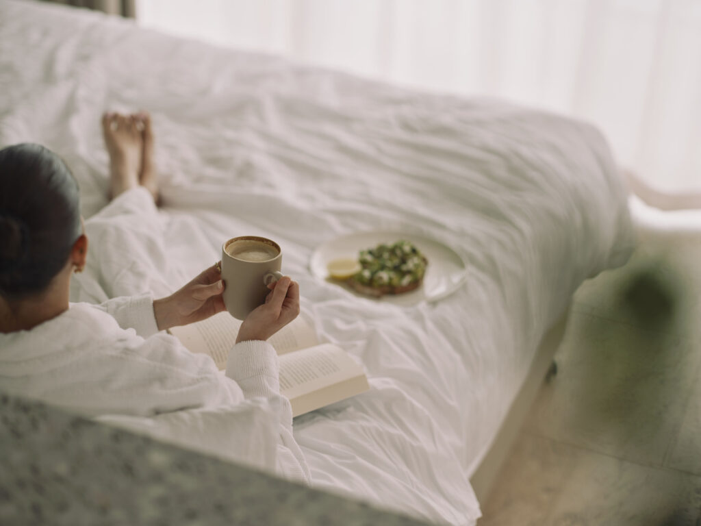 Girl laying in Ardo hotel room bed, wrapped in robe with coffee in hand and book by her side.